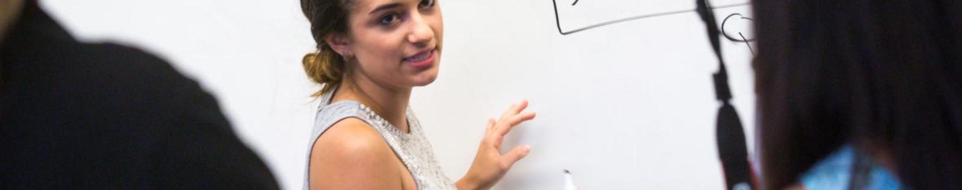 Woman standing in front of whiteboard holding a marker and speaking into a camera.